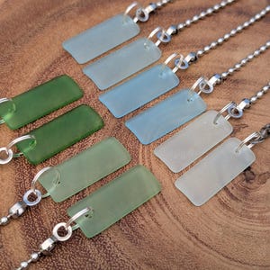 May include: A collection of rectangular sea glass pendants in various shades of green, blue, and white, each attached to a silver-toned metal ball chain by a small jump ring. The glass pieces have a smooth, frosted, and matte finish.