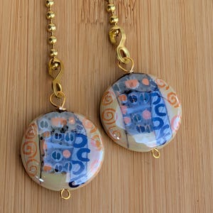 May include: Two decorative ceiling fan pulls. Each pull features a round, glazed ceramic pendant with a colorful abstract design in blue, orange, and beige. The pendants are suspended from gold-colored ball chains.