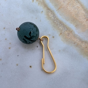 May include: A small, dark green bead with a black design is attached to a gold-coloured clasp. The bead is round and has a textured surface. The clasp is a simple, curved design.