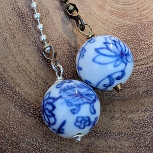 May include: Two white and blue floral patterned ceiling fan pull chains. One chain is silver, the other is bronze. The round pulls have a blue floral design. The background is a brown, textured surface.