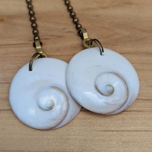May include: Two white shell pendants with a spiral design, each suspended from a bronze-colored chain. The pendants are round with a smooth, glossy finish and a subtle brown hue in the spiral center. The background is a light brown wooden surface.