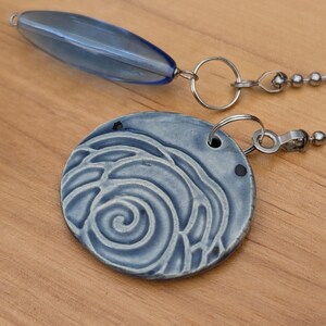 May include: A ceiling fan pull chain featuring a round, light blue ceramic pendant with a raised spiral pattern and a separate, elongated, translucent blue glass bead, both attached to silver-toned metal ball chains with small circular jump rings.