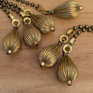 May include: Close-up of several brass-colored, ribbed teardrop-shaped pull chains for a light fixture. The chains are attached to a matching brass-colored ball chain and are arranged on a wooden surface.