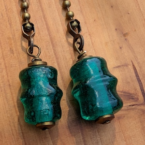 May include: Two decorative, emerald green glass light pull chains with brass-colored hardware. The glass pulls have a unique, textured shape. The chains are suspended against a wooden background.