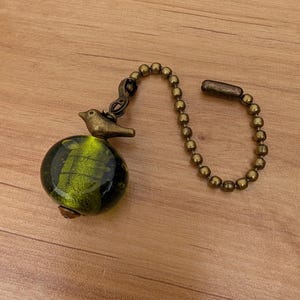 May include: A ceiling fan pull chain featuring a round, translucent green glass bead with internal gold-toned flecks. A small, antique brass-colored metal bird figurine sits atop the glass bead, connected to a matching brass-toned ball chain.