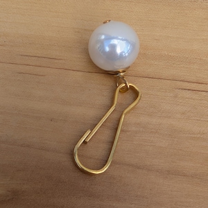 May include: A white pearl charm with a gold metal hook. The pearl is round and has a smooth surface. The hook is a simple design with a loop at the top.