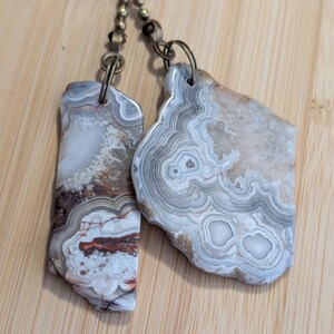 May include: Two irregular, flat agate stone pendants featuring intricate, concentric banding in shades of white, gray, and reddish-brown. Each polished stone is suspended from a bronze-toned metal jump ring attached to a matching ball chain.