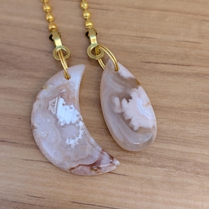 May include: Two light-colored, semi-translucent stone pendants attached to gold-toned metal ball chains. One pendant is shaped like a crescent moon, and the other is shaped like a teardrop. Both stones feature swirling white and beige patterns.