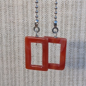May include: Two rectangular, translucent orange resin pendants suspended from silver-toned metal ball chains. Each pendant features a smooth, glossy finish with a hollow center and a small metal jump ring connector at the top for attachment.