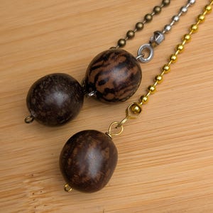 May include: Three ceiling fan pulls featuring smooth, dark brown, oval-shaped wooden beads with natural grain patterns. Each bead is attached to a different metal ball chain: one antique bronze, one silver-toned, and one gold-toned metal chain.