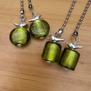 May include: Four ceiling fan pulls featuring silver-toned metal bird charms attached to metallic ball chains. Two pulls have round, textured olive-green glass beads, while the other two have cylindrical, textured olive-green glass beads.