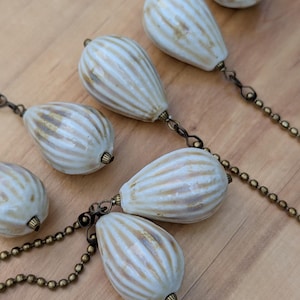 May include: A collection of teardrop-shaped ceramic beads with vertical ridges and a cream-colored glaze featuring brown accents. The beads are connected by antique brass-toned metal ball chains and small jump rings, creating a linked arrangement.