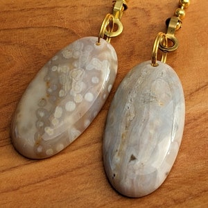 May include: Two oval-shaped, polished gemstones with a light gray and beige color palette. Each stone is attached to a gold-colored chain with a small clasp. The gemstones have unique patterns and textures.