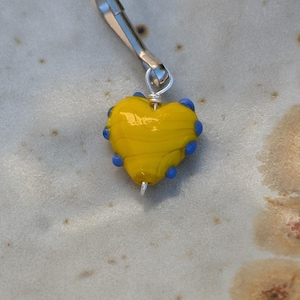 May include: A yellow glass heart charm with blue accents attached to a silver clip.