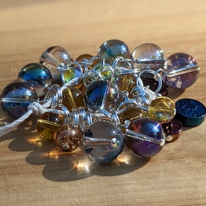 May include: A collection of iridescent glass beads in various sizes and colors, including gold, blue, and purple, strung together with silver wire. A few small, dark blue, and brown decorative beads are also present. The beads are on a light brown wooden surface.