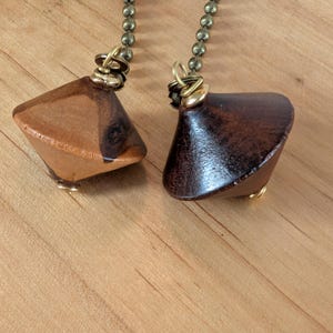 May include: Two ceiling fan pull chains featuring polished wooden finials. One finial is a light brown wood with a dark grain pattern, while the other is a solid dark brown wood. Both are attached to metallic ball chains with gold-toned wire loops.