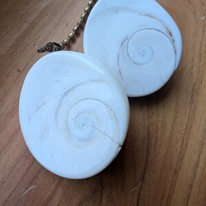 May include: Two white, circular shiva eye shell pendants featuring a natural spiral pattern on the front surface. Each pendant is attached to a metallic ball chain connector, showcasing a smooth, polished finish with subtle beige-toned lines.