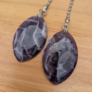 May include: Two marquise-shaped pendants made of purple and white banded amethyst stone. Each pendant features a polished, smooth surface with natural veining and is attached to a silver-toned metal ball chain connector via a small metal jump ring.