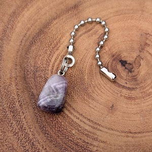 May include: A purple and gray stone pendant with a silver-colored chain and clasp. The pendant is a rounded, polished stone with a mix of colors. The chain is made of small, connected metal balls. The pendant hangs on a wooden surface.