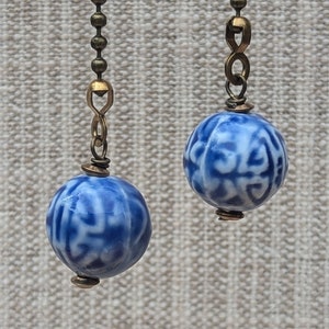 May include: Two blue and white ceramic ball fan pulls with a detailed pattern. The pulls are attached to a brass-colored chain. The background is a neutral, textured fabric.