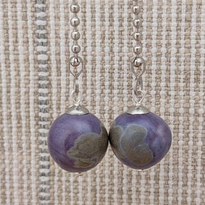 May include: A pair of drop earrings featuring spherical purple beads with mottled grey-brown patches. Each bead is attached to a silver-toned metal ball chain by a small silver jump ring and a polished silver-toned metal cap at the top of the bead.