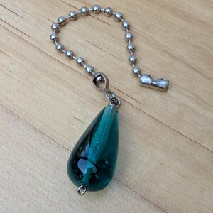 May include: A teardrop-shaped pendant made of translucent teal glass, featuring a small metal loop at the bottom. The pendant is attached to a silver-toned metal ball chain connector, which includes a small metal clasp at the end of the chain.