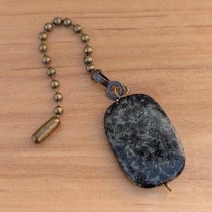 May include: A dark green and black stone pendant with a brass-colored chain and clasp. The pendant is rectangular with rounded edges. The chain is made of small, round beads. The background is a light brown wooden surface.