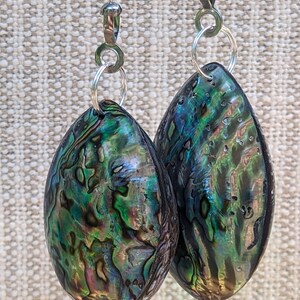 May include: A pair of teardrop-shaped earrings featuring iridescent abalone shell in shades of deep green, blue, and purple. Each shell is suspended from a silver-toned metal jump ring and a silver-toned ball chain connector, showcasing a natural, polished, and multi-colored surface texture.