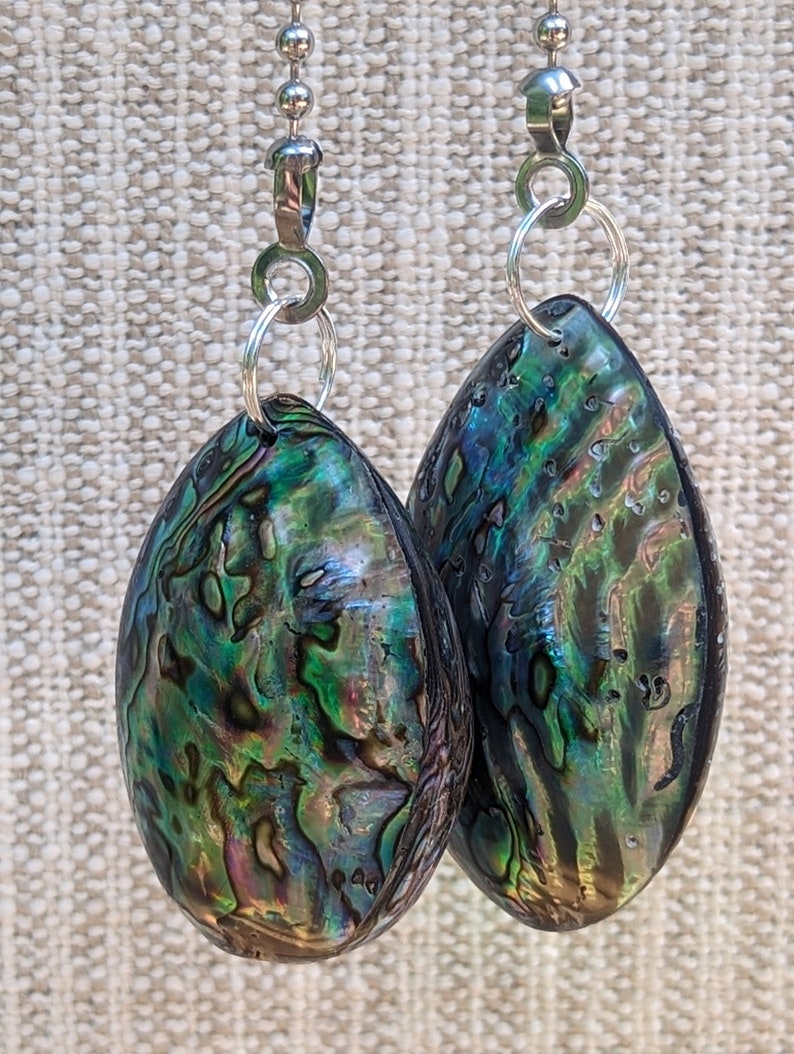 May include: A pair of teardrop-shaped earrings featuring iridescent abalone shell in shades of deep green, blue, and purple. Each shell is suspended from a silver-toned metal jump ring and a silver-toned ball chain connector, showcasing a natural, polished, and multi-colored surface texture.
