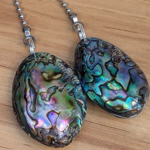 May include: Two oval-shaped abalone shell pendants attached to silver-toned metal ball chains. The shells feature a natural iridescent surface with swirling patterns of deep blue, vibrant green, purple, and black hues, polished to a smooth finish.