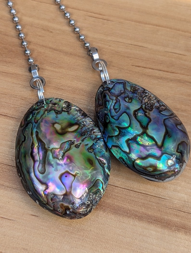 May include: Two oval-shaped abalone shell pendants attached to silver-toned metal ball chains. The shells feature a natural iridescent surface with swirling patterns of deep blue, vibrant green, purple, and black hues, polished to a smooth finish.