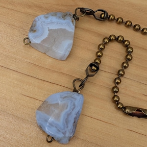 May include: Two blue and beige agate stone pendants connected by antique brass chains and a beaded chain. The stones have a natural, layered appearance. The background is a light brown wooden surface.