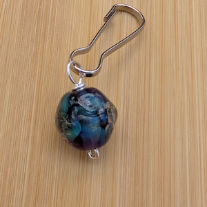 May include: A silver metal clip with a round, dark blue and purple glass bead with a floral design. The bead is attached to the clip with a silver wire loop.