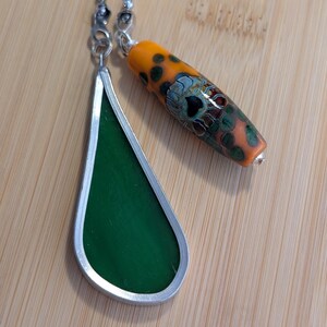 May include: A handmade necklace featuring two pendants. One is a teardrop-shaped, green glass pendant with a silver-colored metal frame. The other is an orange and green glass bead with a floral design. The necklace has a silver-colored chain.