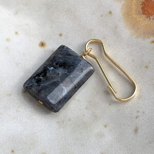 May include: A rectangular dark gray stone pendant with a polished, mottled surface featuring subtle blue iridescent flashes, attached to a simple gold-toned metal hook clasp. The stone has a smooth, flat finish and is secured by a small gold pin.