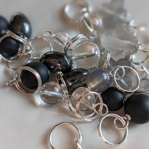 May include: A close-up of a collection of black, gray, and clear gemstone beads with silver jump rings. The beads are arranged in a random pattern and are all different sizes.