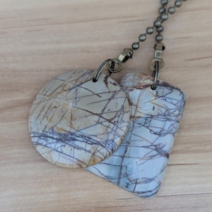May include: Two stone pendant necklaces. One pendant is a circle, the other is a modified rectangle. Both pendants are beige with dark brown and blue veining. The necklaces have a bronze-colored chain.