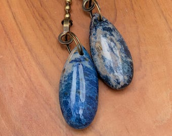 May include: Two polished, teardrop-shaped blue and white stone pendants attached to antique brass-colored metal ball chains. The stones feature marbled patterns with deep blue, light blue, and white veins, each secured by a metal jump ring.