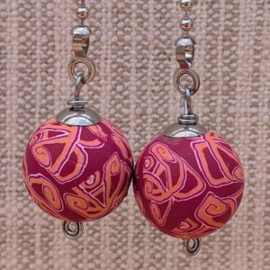 May include: Two decorative ceiling fan pull chains. Each chain features a round, maroon-colored ball with an orange swirl design. The balls are attached to a silver chain with a decorative metal clasp.