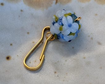 May include: A gold-toned metal hook clasp attached to a spherical glass bead. The bead features a light blue floral pattern with small white petals, dark blue centers, and green leaf accents embedded within the translucent glass surface.