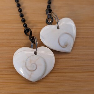 May include: Two heart-shaped pendants with a spiral design, suspended from black beaded chains. The pendants are white with a natural, swirling pattern. The background is a light brown wooden surface.