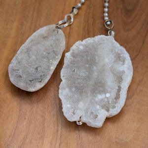 May include: Two irregular, off-white geode slices attached to silver-toned ball chains. The larger geode is roughly circular with a crystalline, textured surface, while the smaller one is elongated and oval-shaped with similar mineral formations.