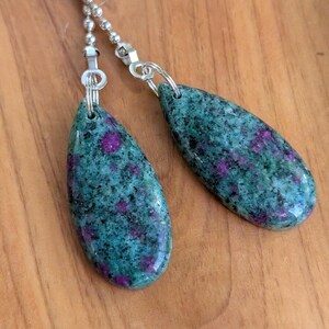 May include: Two teardrop-shaped pendants made of green zoisite stone with embedded magenta ruby crystals. Each pendant is attached to a silver-toned metal ball chain connector by a small circular jump ring, featuring a polished, smooth surface.