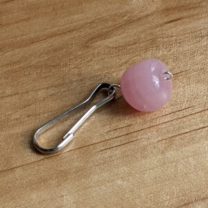 May include: A silver-toned metal lobster clasp attached to a single, smooth, round, light pink bead. The bead has a glossy, semi-translucent finish and is connected to the clasp by a small silver-colored metal jump ring.