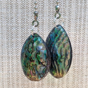 May include: Two teardrop-shaped abalone shell pendants featuring iridescent swirls of green, blue, and purple. Each pendant is attached to a silver-toned metal jump ring and a silver ball chain, showcasing a polished, smooth, and reflective surface.