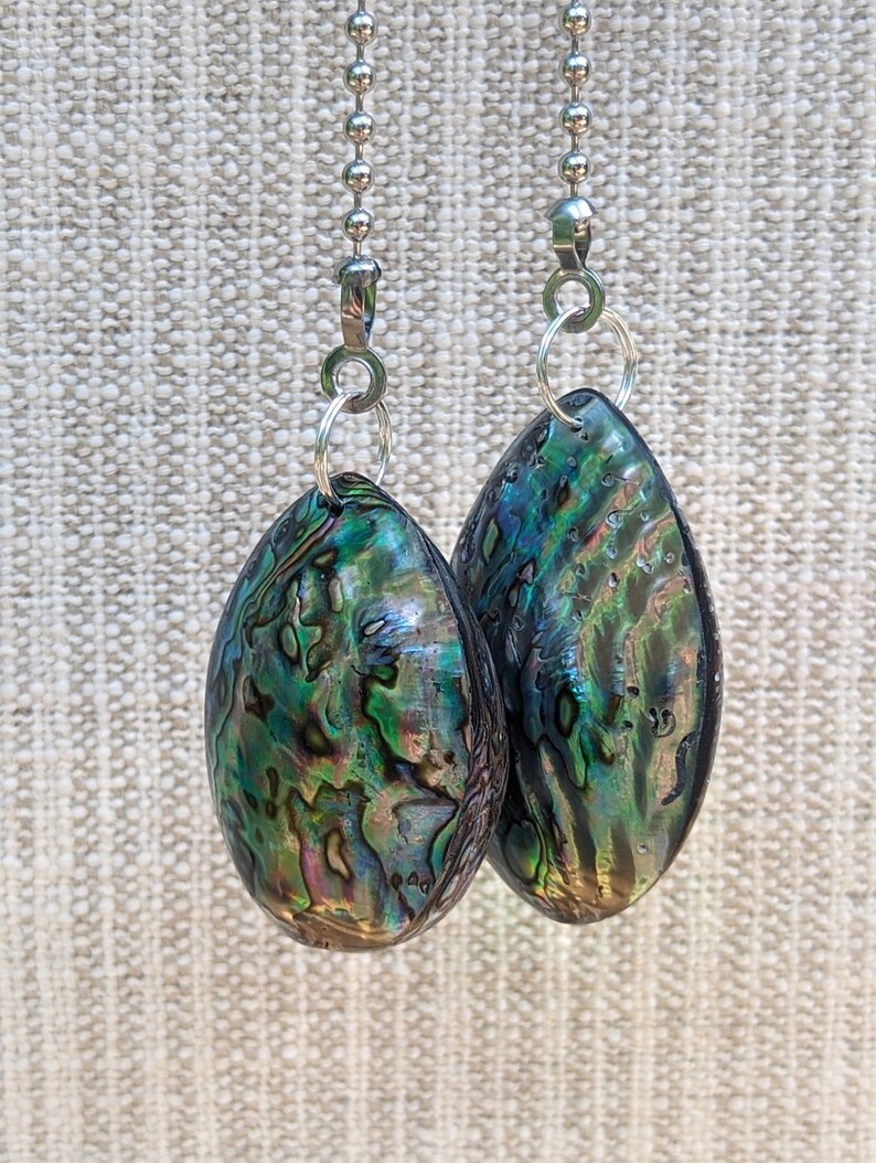 May include: Two teardrop-shaped abalone shell pendants featuring iridescent swirls of green, blue, and purple. Each pendant is attached to a silver-toned metal jump ring and a silver ball chain, showcasing a polished, smooth, and reflective surface.