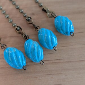 May include: Close-up of four turquoise-colored, spiral-shaped beads hanging from antique bronze-colored chains. The beads have a glossy finish and are connected to the chains with small metal loops. The background is a light-colored wooden surface.