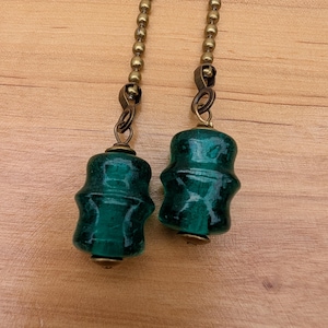 May include: Two dark green glass beads attached to antique brass ball chains. Each bead features a distinct, multi-tiered cylindrical shape with a smooth, glossy finish. The metal chains are connected to the top of each glass piece with small brass loops.