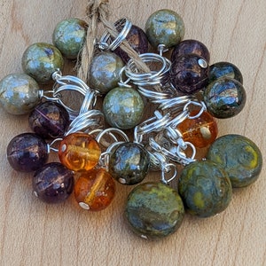 May include: A collection of colorful glass beads with silver wire loops. The beads are in shades of green, purple, amber, and brown.