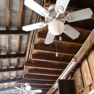 May include: Two white ceiling fans with five blades each and a central globe light fixture. Each fan features a pull chain for operation, mounted against a dark brown wooden beamed ceiling structure with visible rafters and wooden planks.